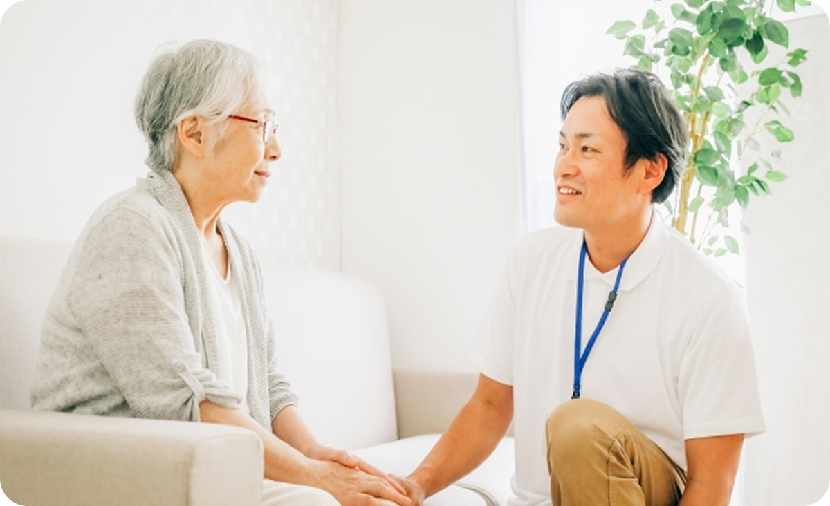 ソファに座り、笑顔で向き合いながら高齢女性の手を優しく握って寄り添う男性スタッフの様子。